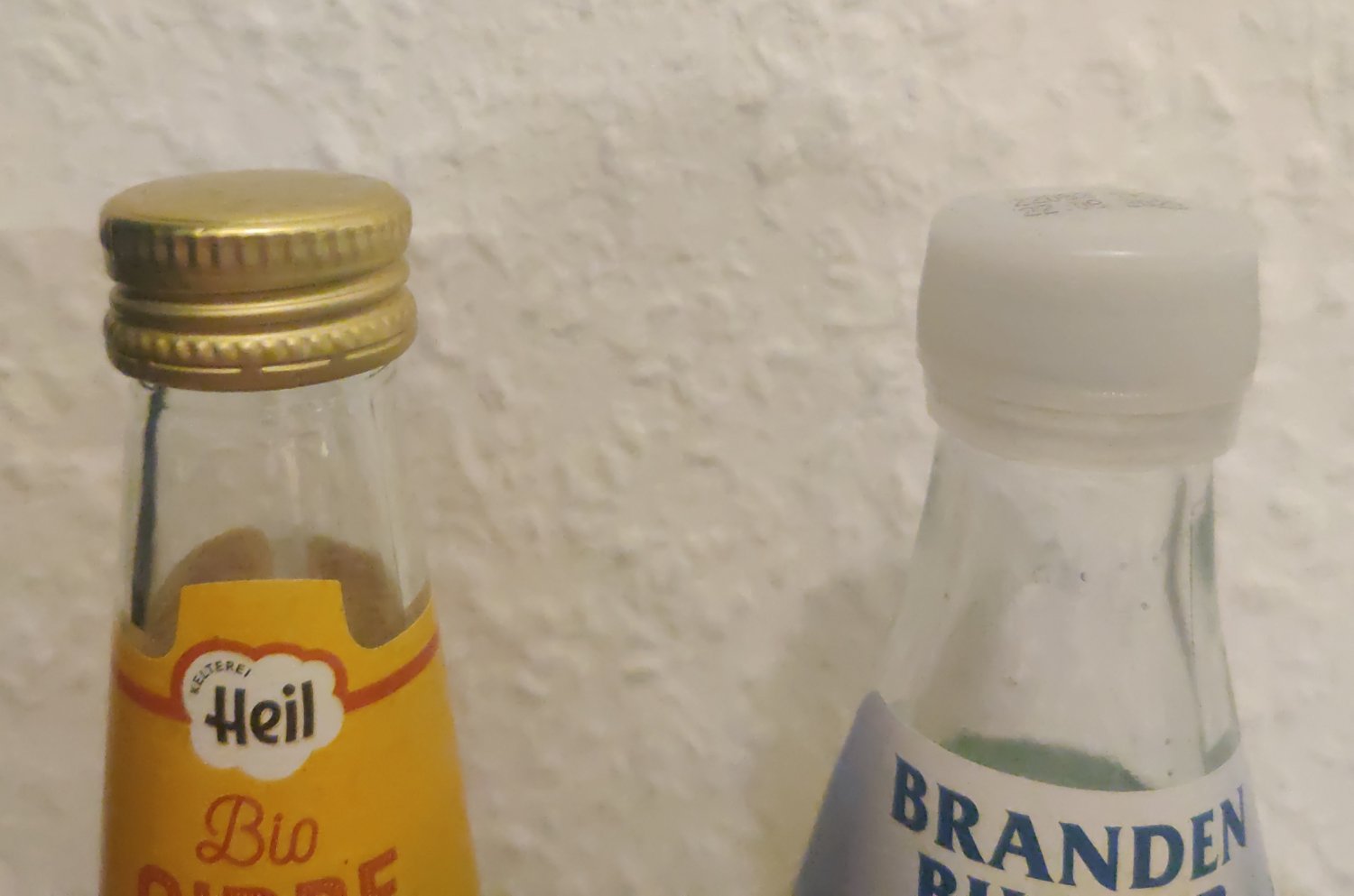 A photo of two glass bottles next to each other. The left has a brass-colored metal cap screwed on, the right a white plastic one.