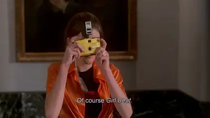 Screenshot from the movie Kids in the Hall: Brain Candy (1996), a young journalist taking a photograph during a small press conference, subtitle has part of the line thanking the few press attendees: "Of course Girl Beat."