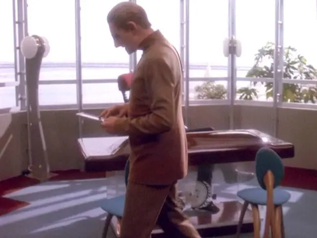 A screencap from Star Trek: Deep Space 9, depicting Constable Odo walking in front of a desk with spindly bulbous chairs.