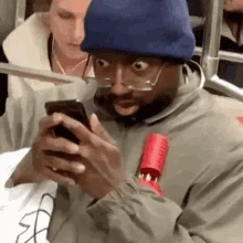 A black man with his glasses pulled down to the tip of his nose, looks down at his cellphone in shock/horror/disbelief with wide open eyes and mouth
