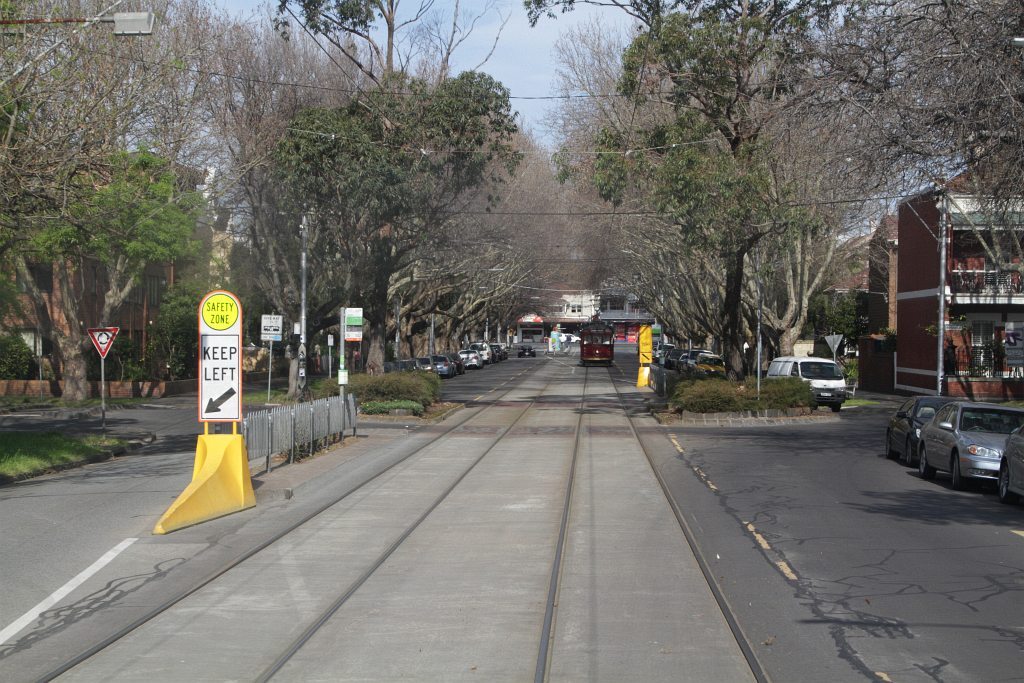 photo of a melbourne tram stop in a leafy street, where the passenger boarding/alighting area is between the car lane and the tram track, protected from cars by a solid concrete block in an elongated tetrahedron shape painted yellow