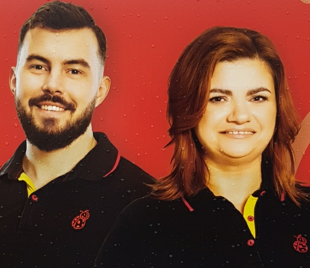 A photo of two employees, male and female. Man is smiling nir ally, while woman is smiling in a painful manner.
